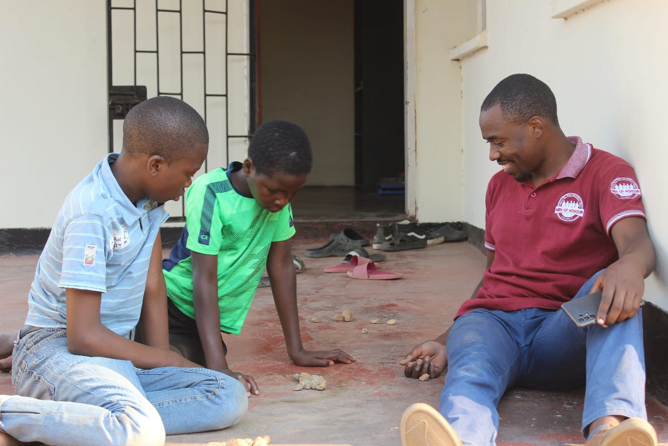 An older Zambian male engaging with two Zambian boys to look at rocks on the floor